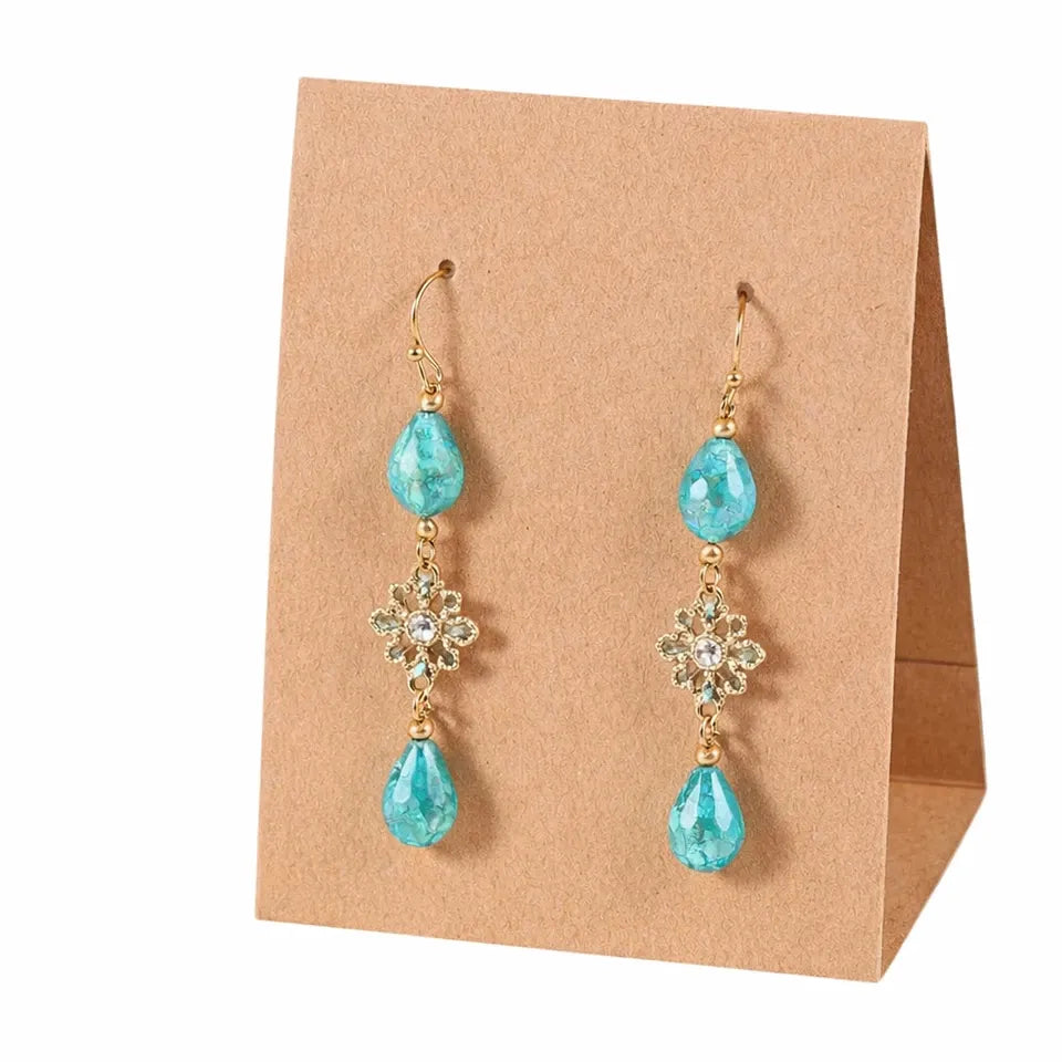 A pair of gold-tone drop earrings with turquoise blue teardrop beads and clear rhinestone floral connectors, displayed on a brown kraft paper stand against a white background.