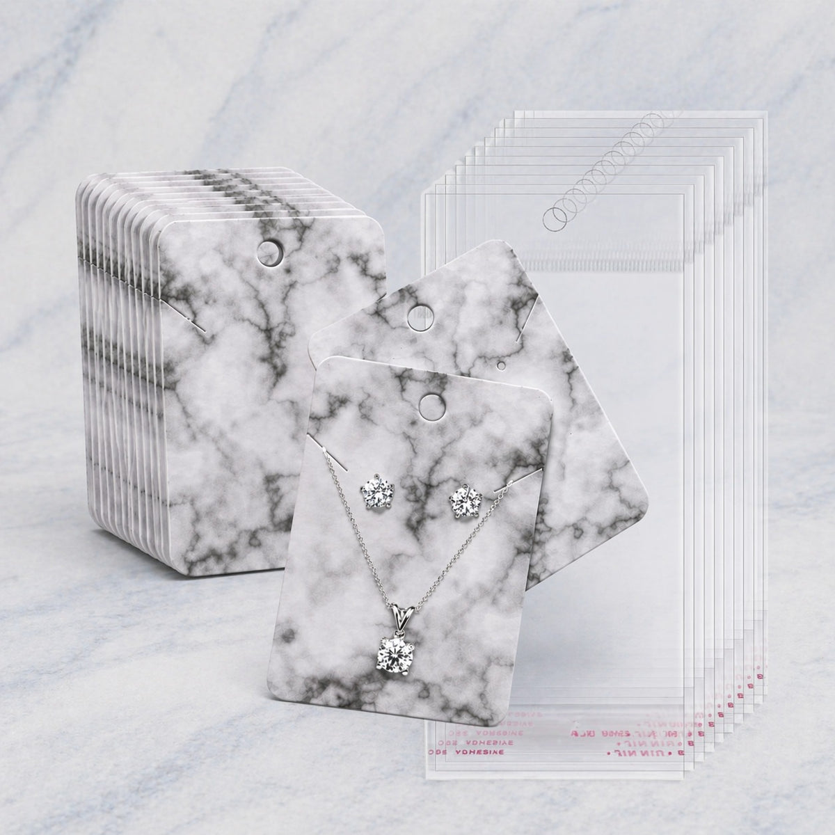 A set of jewelry display cards with white and gray marble-patterned backing, shown with a necklace and matching stud earrings displayed on one card, alongside a stack of blank cards and a stack of clear cards on a light marble surface.