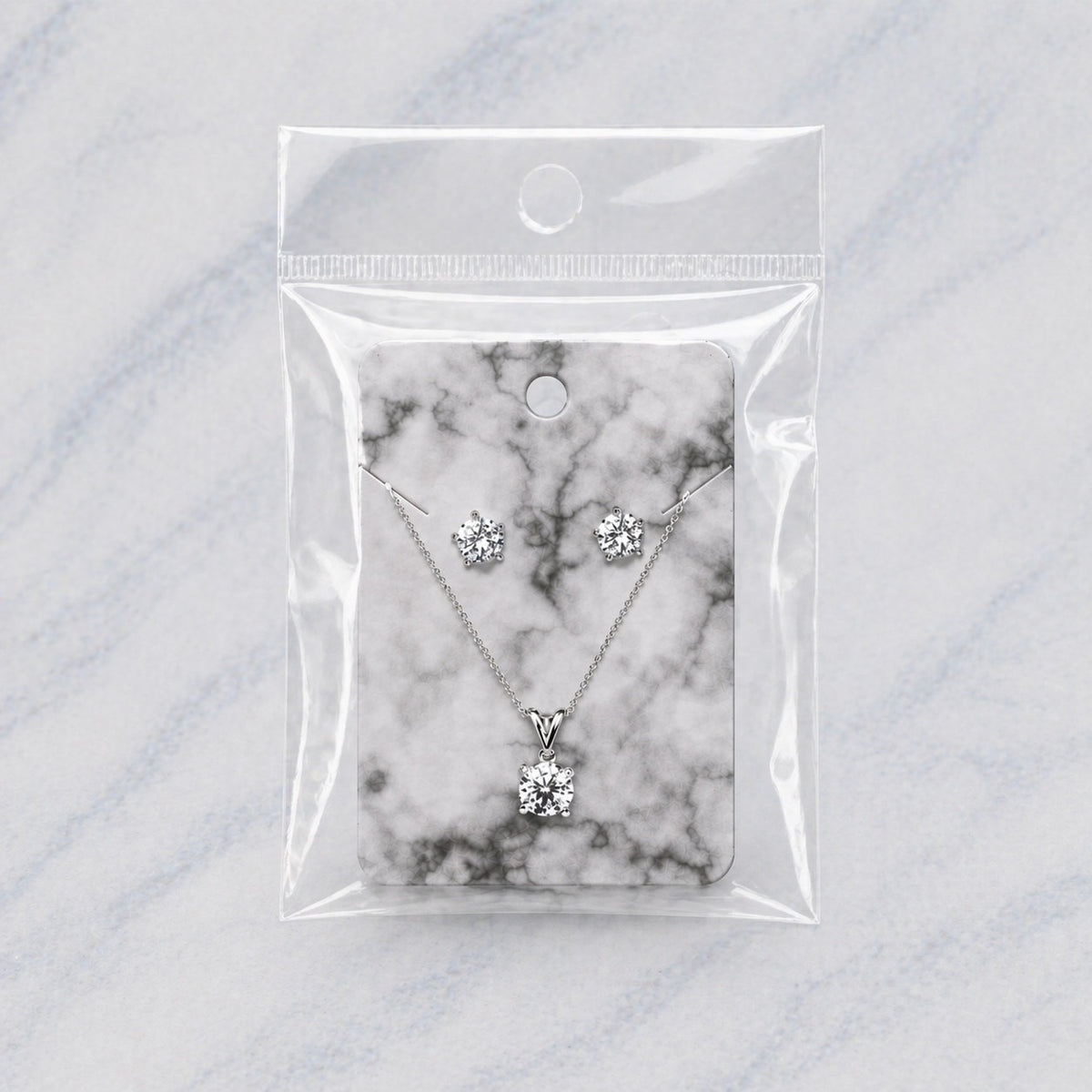 Silver necklace and matching stud earrings displayed on a black-and-grey marble card inside a clear plastic hanging bag, set on a light marble surface.