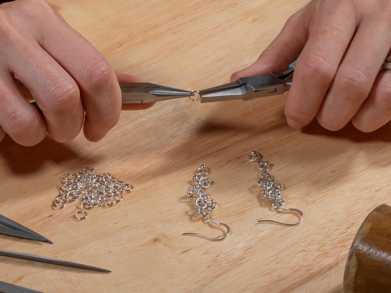 Close-up of hands using silver-toned pliers to join small chain links, with finished silver dangle earrings and loose jump rings on a light wood surface.
