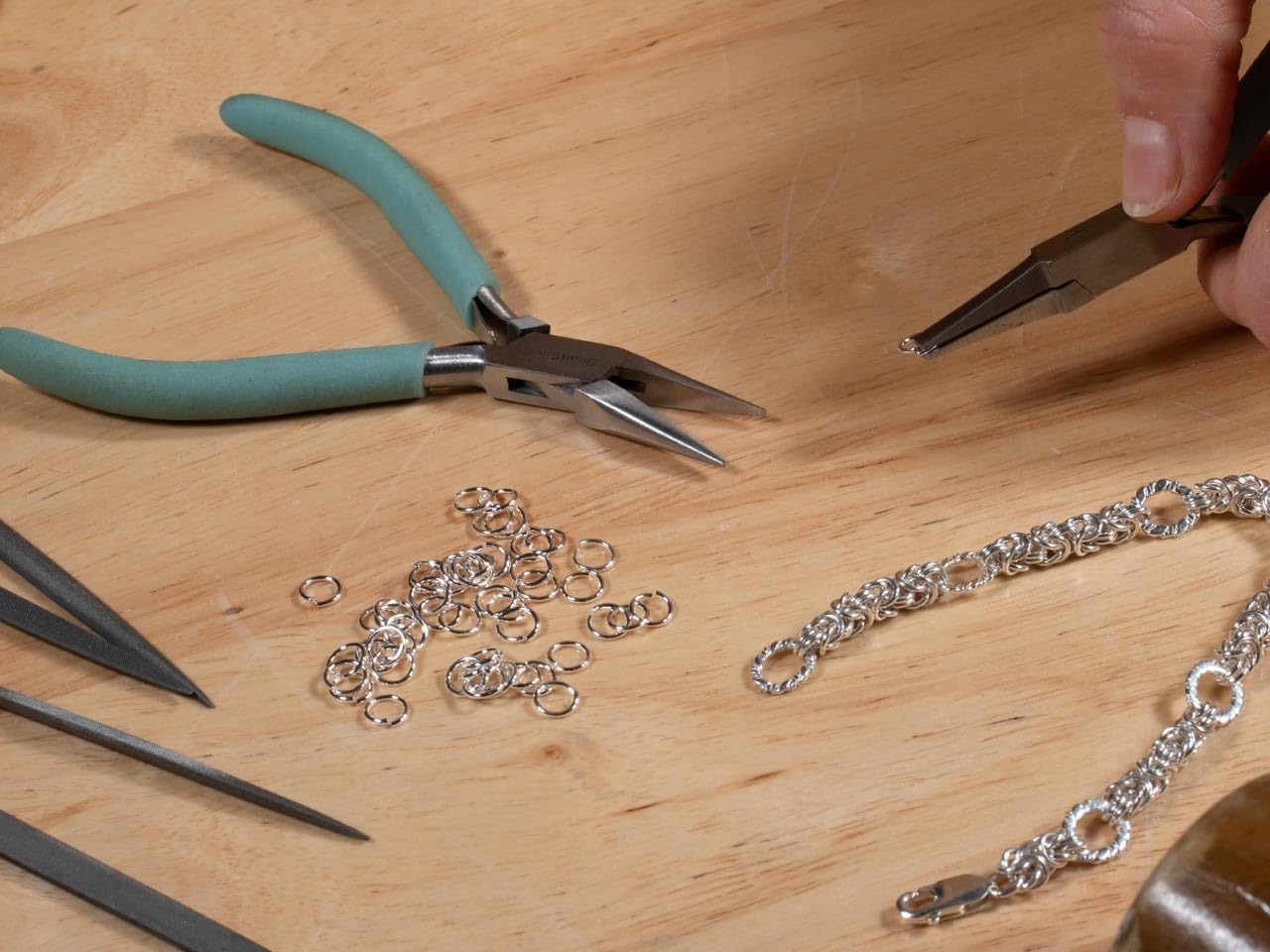 A close-up of jewelry-making tools on a wooden surface, including teal-handled pliers, small silver jump rings, and a silver chain, with a hand holding a metal tool.